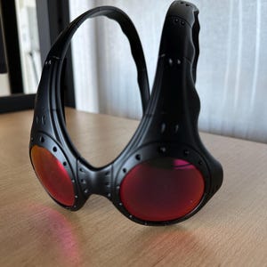May include: Black sunglasses with red lenses. The sunglasses have a unique, wraparound design with a thick frame and visible rivets. The lenses are a vibrant red color, contrasting with the black frame. The overall style is modern and sporty.