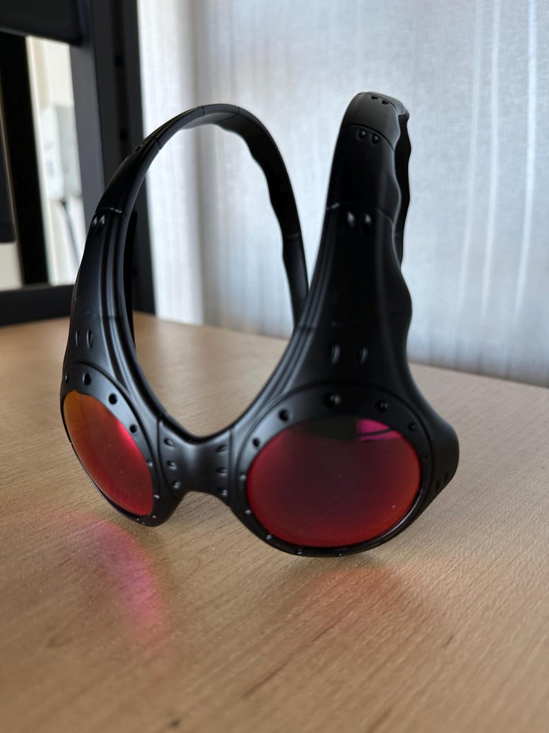 May include: Black sunglasses with red lenses. The sunglasses have a unique, wraparound design with a thick frame and visible rivets. The lenses are a vibrant red color, contrasting with the black frame. The overall style is modern and sporty.