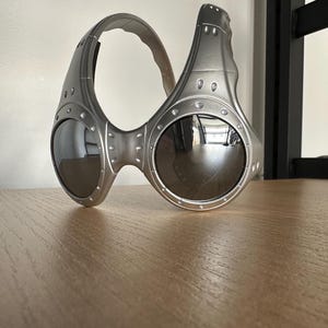May include: Silver sunglasses with a futuristic design and dark lenses. The glasses have a unique, wraparound style with a thick frame and visible rivets. The lenses are round and reflective, and the frame has a matte finish.