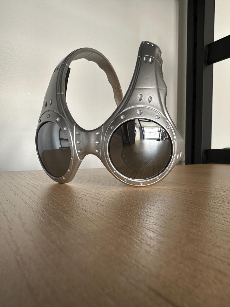 May include: Silver sunglasses with a futuristic design and dark lenses. The glasses have a unique, wraparound style with a thick frame and visible rivets. The lenses are round and reflective, and the frame has a matte finish.