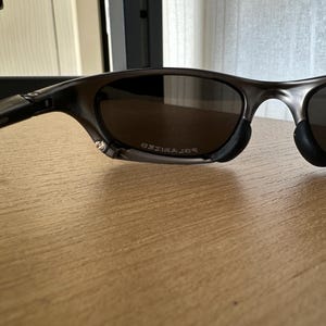 May include: Dark grey sunglasses with polarised lenses. The sunglasses have a sleek, modern design with a curved frame and black nose pads. The word "POLARIZED" is visible on the lens.
