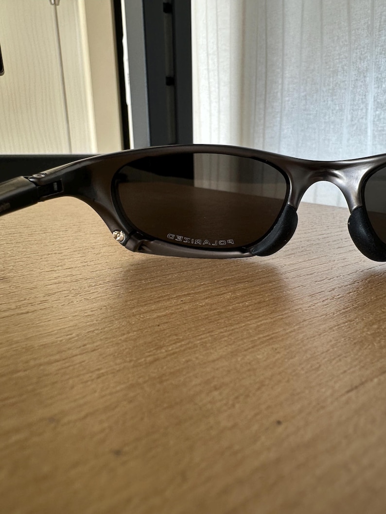 May include: Dark grey sunglasses with polarised lenses. The sunglasses have a sleek, modern design with a curved frame and black nose pads. The word "POLARIZED" is visible on the lens.