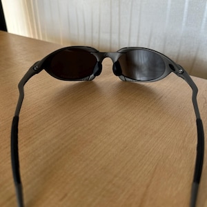 May include: Dark gray sunglasses with oval lenses and a thin, curved frame. The lenses are a dark tint, and the arms have black rubberized grips for comfort. The sunglasses are designed for a sleek, modern look.