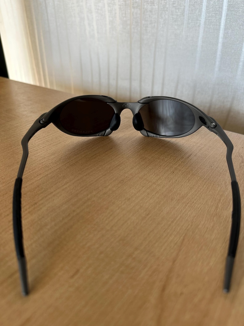 May include: Dark gray sunglasses with oval lenses and a thin, curved frame. The lenses are a dark tint, and the arms have black rubberized grips for comfort. The sunglasses are designed for a sleek, modern look.