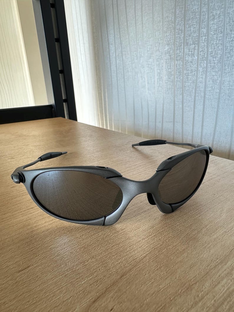 May include: Dark gray sunglasses with oval lenses and a wraparound design. The frames are a matte finish, and the lenses have a mirrored appearance. The sunglasses are resting on a light-colored wooden surface.