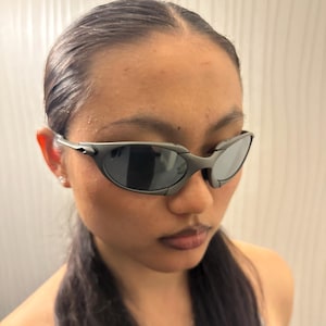 May include: Dark gray sunglasses with a curved, wraparound design. The lenses are dark, and the frames have a matte finish. The sunglasses are a sporty style, suitable for outdoor activities.