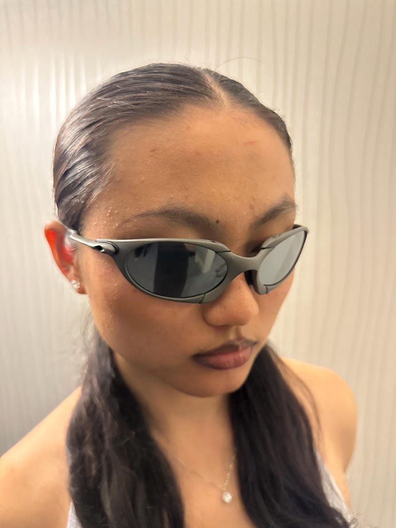 May include: Dark gray sunglasses with a curved, wraparound design. The lenses are dark, and the frames have a matte finish. The sunglasses are a sporty style, suitable for outdoor activities.