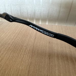 Pu&ograve; includere: Primo piano di un'asta di occhiali nera con la scritta "MADMAN" stampata in bianco. L'asta ha un design curvo ed &egrave; appoggiata su una superficie di legno marrone chiaro e uno sfondo a righe bianche e beige.