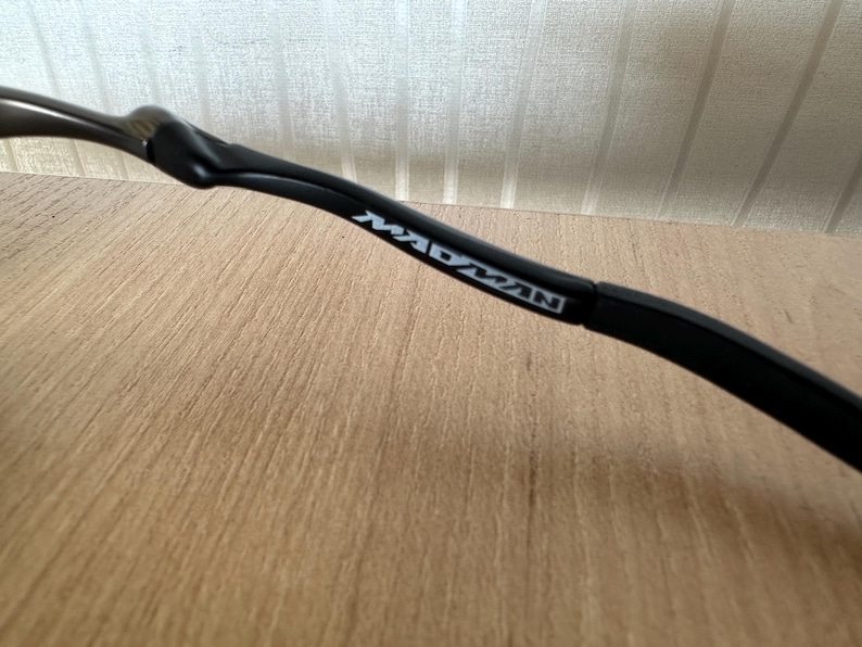 Pu&ograve; includere: Primo piano di un'asta di occhiali nera con la scritta "MADMAN" stampata in bianco. L'asta ha un design curvo ed &egrave; appoggiata su una superficie di legno marrone chiaro e uno sfondo a righe bianche e beige.