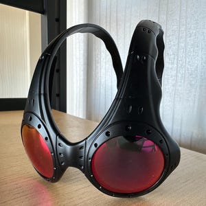 May include: Black sunglasses with red lenses. The sunglasses have a unique, wraparound design with a thick frame and visible rivets. The lenses are circular and the frame is matte black.