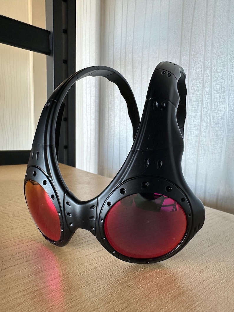 May include: Black sunglasses with red lenses. The sunglasses have a unique, wraparound design with a thick frame and visible rivets. The lenses are circular and the frame is matte black.