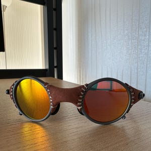 May include: Sunglasses with a brown leather bridge and temples, featuring round lenses with a gradient of yellow to orange and red. The frames are black with white stitching details.