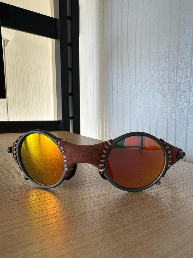 May include: Sunglasses with a brown leather bridge and temples, featuring round lenses with a gradient of yellow to orange and red. The frames are black with white stitching details.