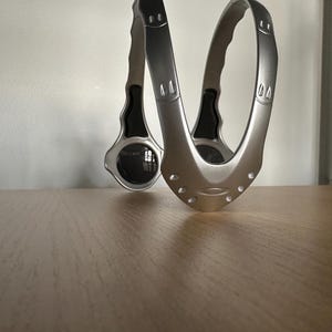 May include: A silver-coloured, curved metal object with a handle on each end. The object has a series of small holes and a circular element on one side. The design is modern and sleek, with a metallic finish.