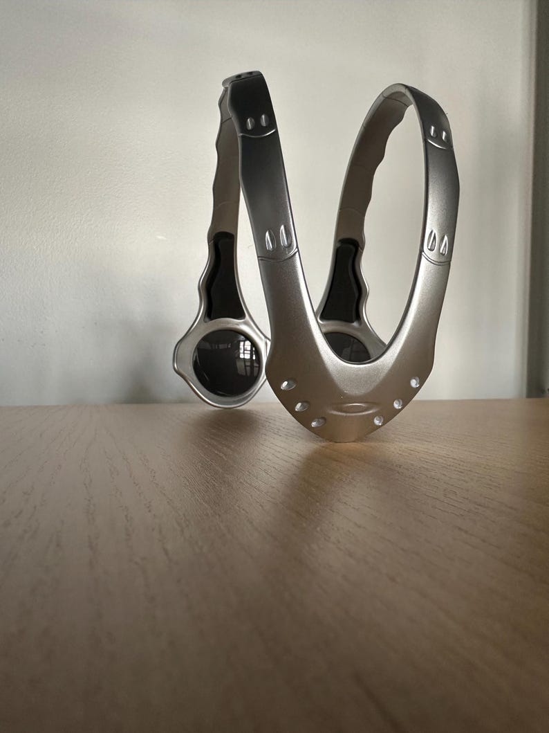 May include: A silver-coloured, curved metal object with a handle on each end. The object has a series of small holes and a circular element on one side. The design is modern and sleek, with a metallic finish.