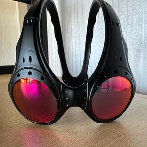 May include: Black goggles with red lenses. The goggles have a unique design with a bridge and two curved arms. The lenses are round and reflect a pink hue. The frame has a riveted appearance.