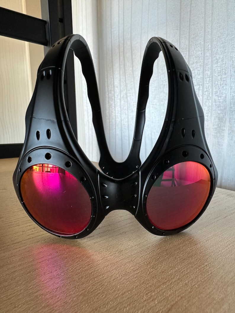 May include: Black goggles with red lenses. The goggles have a unique design with a bridge and two curved arms. The lenses are round and reflect a pink hue. The frame has a riveted appearance.