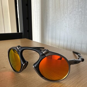 Pu&ograve; includere: Un paio di occhiali da sole con montatura nera e lenti arancioni. Il design &egrave; curvo e moderno. Le lenti riflettono una tonalit&agrave; arancione brillante. Gli occhiali sono su una superficie di legno.