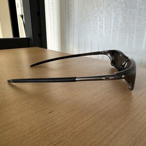 May include: Dark grey sunglasses with a streamlined design. The sunglasses feature a curved frame and dark lenses. The arms are thin and extend to the temples. The sunglasses are resting on a light brown wooden surface.