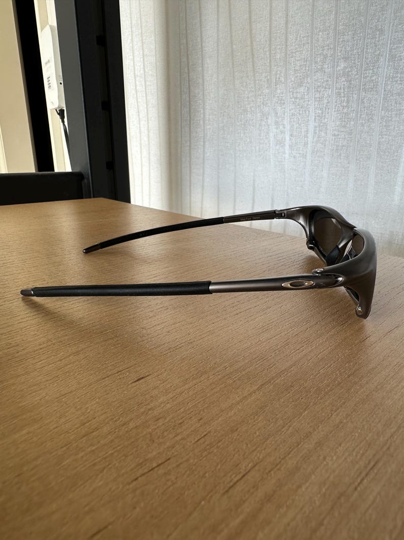 May include: Dark grey sunglasses with a streamlined design. The sunglasses feature a curved frame and dark lenses. The arms are thin and extend to the temples. The sunglasses are resting on a light brown wooden surface.