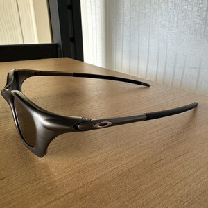 May include: Dark grey sunglasses with a sleek, modern design. The lenses are a dark tint, and the frames have a matte finish. The temples are thin and curve around the ears. The sunglasses are resting on a wooden surface.