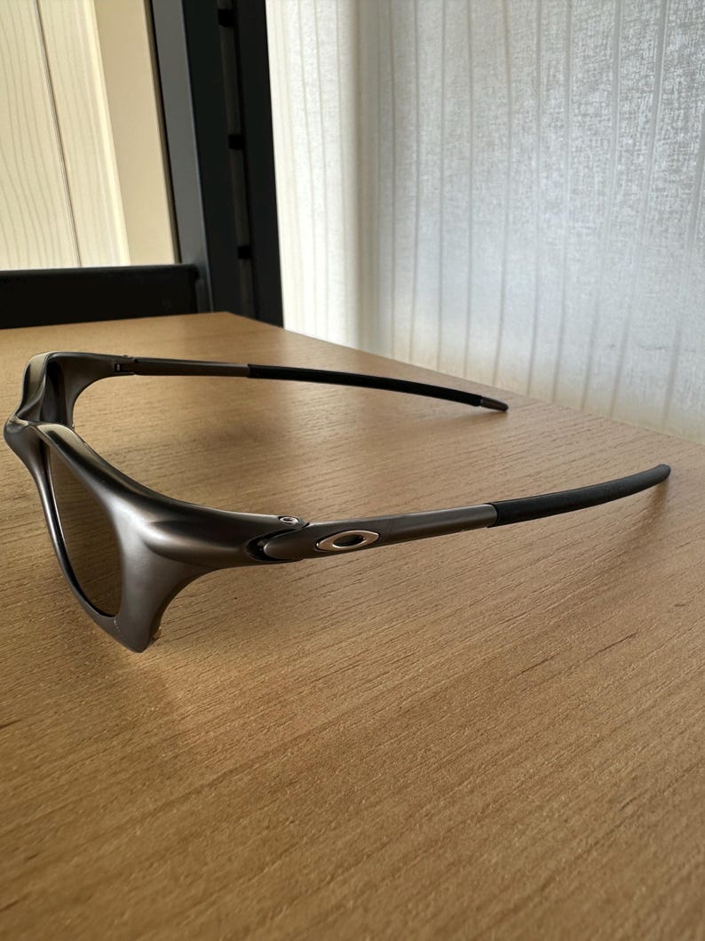 May include: Dark grey sunglasses with a sleek, modern design. The lenses are a dark tint, and the frames have a matte finish. The temples are thin and curve around the ears. The sunglasses are resting on a wooden surface.