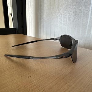 Pode incluir: &Oacute;culos de sol cinza escuro com um design elegante e moderno. Os &oacute;culos t&ecirc;m uma arma&ccedil;&atilde;o curva e lentes escuras. As hastes s&atilde;o finas e estendem-se para tr&aacute;s da orelha. O logotipo da Oakley &eacute; vis&iacute;vel na haste.