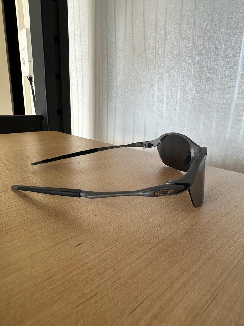 Pode incluir: &Oacute;culos de sol cinza escuro com um design elegante e moderno. Os &oacute;culos t&ecirc;m uma arma&ccedil;&atilde;o curva e lentes escuras. As hastes s&atilde;o finas e estendem-se para tr&aacute;s da orelha. O logotipo da Oakley &eacute; vis&iacute;vel na haste.