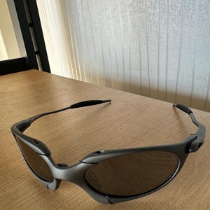 May include: Gray sunglasses with dark lenses rest on a wooden surface. The sunglasses have a sleek, modern design with a curved frame and a black band at the temples. The lenses are oval-shaped.