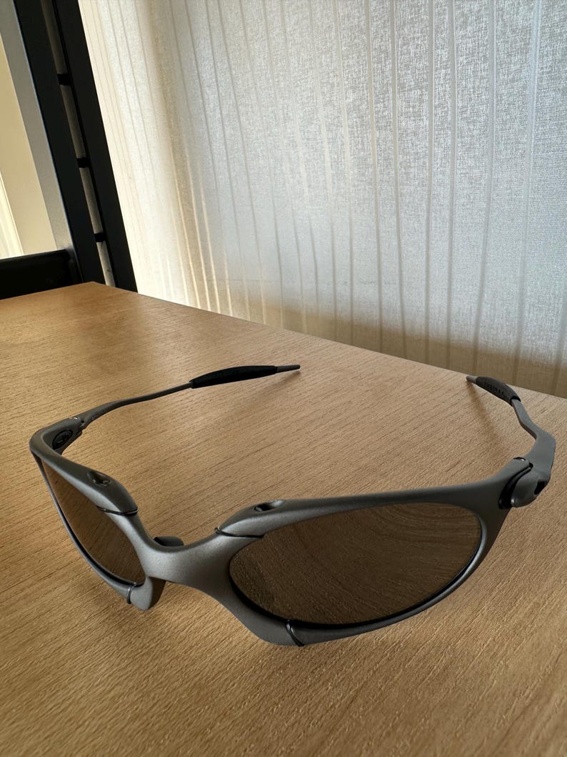 May include: Gray sunglasses with dark lenses rest on a wooden surface. The sunglasses have a sleek, modern design with a curved frame and a black band at the temples. The lenses are oval-shaped.