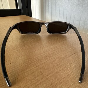 May include: Sunglasses with a dark frame and brown lenses, set on a wooden surface. The glasses have a modern design with curved arms and a metallic finish. The lenses appear to be polarized.