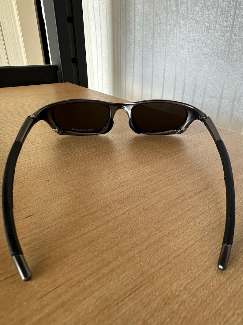 May include: Sunglasses with a dark frame and brown lenses, set on a wooden surface. The glasses have a modern design with curved arms and a metallic finish. The lenses appear to be polarized.
