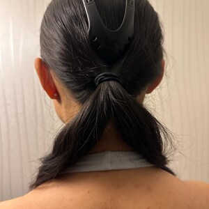 May include: A black hair accessory is used to style dark hair into a ponytail. The accessory is a curved shape with a series of small holes and is positioned at the top of the head. The hair is pulled back and secured with a black elastic band.