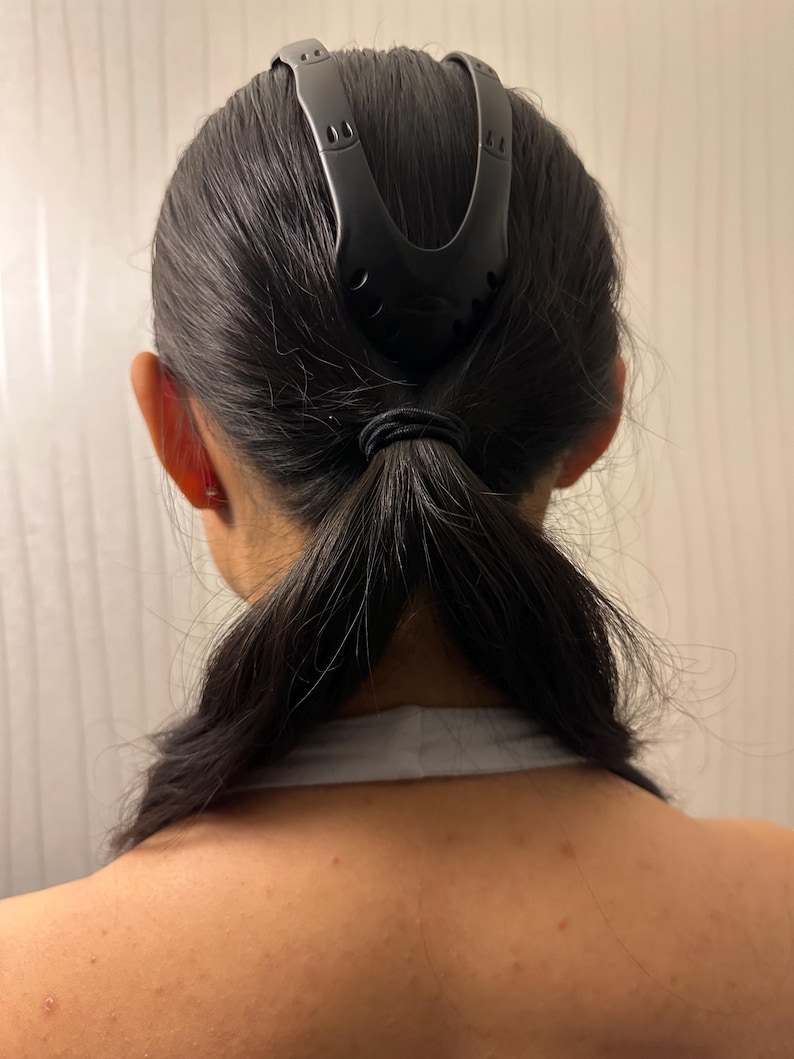 May include: A black hair accessory is used to style dark hair into a ponytail. The accessory is a curved shape with a series of small holes and is positioned at the top of the head. The hair is pulled back and secured with a black elastic band.