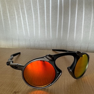Pu&ograve; includere: Occhiali da sole neri e grigi con lenti rotonde tinte di rosso. Gli occhiali hanno un design unico con montatura curva. Le lenti riflettono un colore arancione brillante. Sono appoggiati su una superficie di legno.