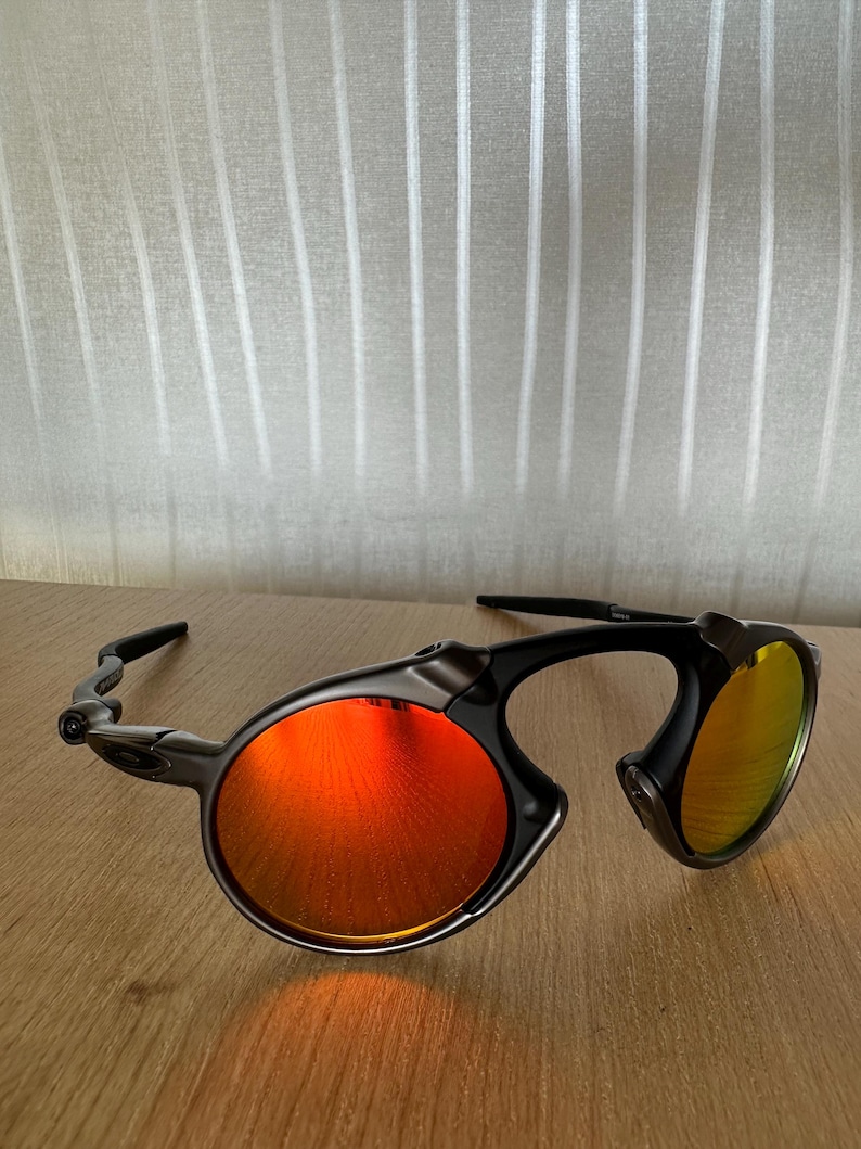 Pu&ograve; includere: Occhiali da sole neri e grigi con lenti rotonde tinte di rosso. Gli occhiali hanno un design unico con montatura curva. Le lenti riflettono un colore arancione brillante. Sono appoggiati su una superficie di legno.
