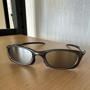 May include: Brown-framed sunglasses with reflective lenses on a wooden surface. The sunglasses have a curved design and a dark, matte finish. The lenses are tinted, providing protection from the sun.