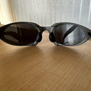 May include: Dark gray sunglasses with a curved frame and dark lenses. The sunglasses have black nose pads and are resting on a wooden surface. The lenses reflect the surrounding environment.