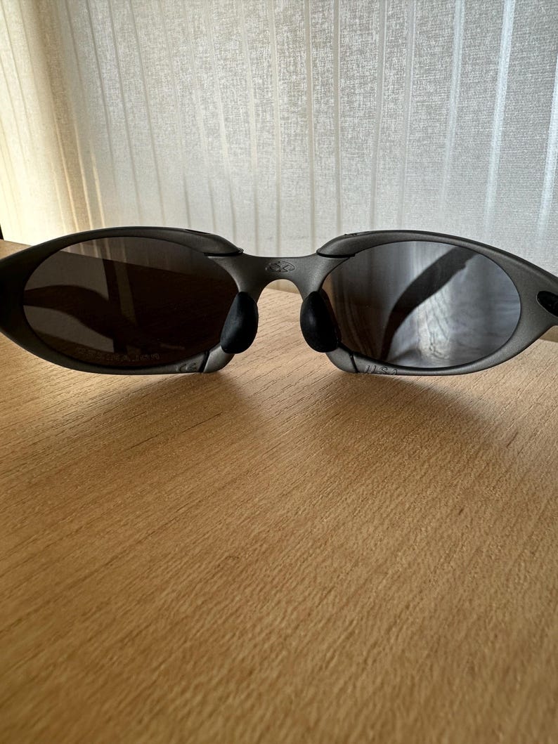 May include: Dark gray sunglasses with a curved frame and dark lenses. The sunglasses have black nose pads and are resting on a wooden surface. The lenses reflect the surrounding environment.