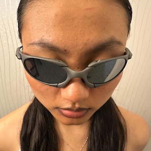 May include: Gray sunglasses with dark lenses. The sunglasses have a wraparound design and a matte finish. The frame is a muted gray color, and the lenses reflect light. The image is a close-up of the sunglasses.
