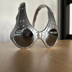 May include: Silver sunglasses with a distinctive, industrial aesthetic. The frames have a riveted design and a curved brow bar. The dark lenses contrast with the metallic finish. A contemporary style of eyewear.