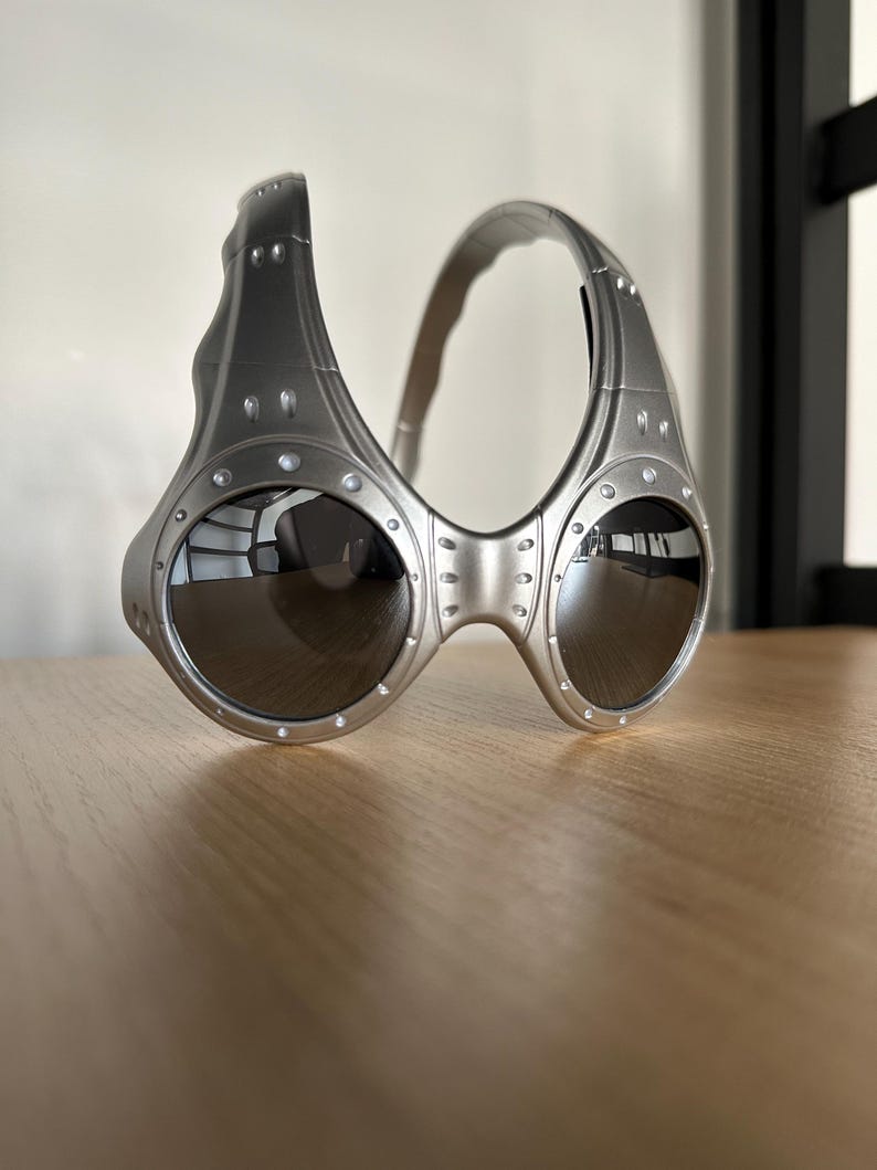 May include: Silver sunglasses with a distinctive, industrial aesthetic. The frames have a riveted design and a curved brow bar. The dark lenses contrast with the metallic finish. A contemporary style of eyewear.