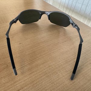 Pu&ograve; includere: Occhiali da sole grigio scuro con un design semi-senza montatura. Le lenti sono di colore verde scuro e le aste hanno impugnature in gomma nera. Gli occhiali da sole sono su una superficie di legno marrone chiaro.