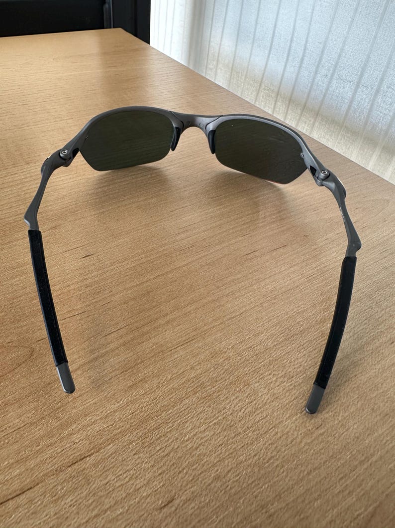 Pu&ograve; includere: Occhiali da sole grigio scuro con un design semi-senza montatura. Le lenti sono di colore verde scuro e le aste hanno impugnature in gomma nera. Gli occhiali da sole sono su una superficie di legno marrone chiaro.