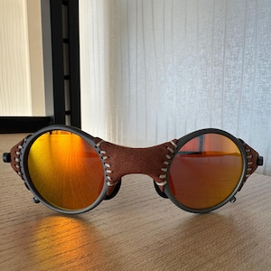 May include: Oval-shaped sunglasses with orange-tinted lenses and a brown leather bridge. The frames are black with a stitched leather detail. The lenses reflect a warm, orange hue, suggesting a sunny environment.