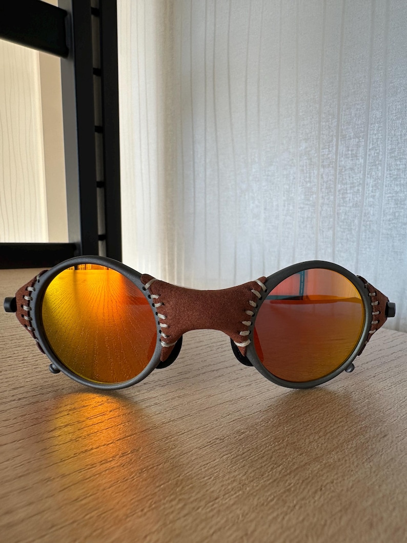 May include: Oval-shaped sunglasses with orange-tinted lenses and a brown leather bridge. The frames are black with a stitched leather detail. The lenses reflect a warm, orange hue, suggesting a sunny environment.