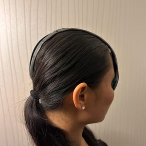 May include: A black headband worn on dark hair. The headband is smooth and curved, and it sits across the forehead. The hair is pulled back into a low ponytail. The person is wearing a small, clear stud earring.