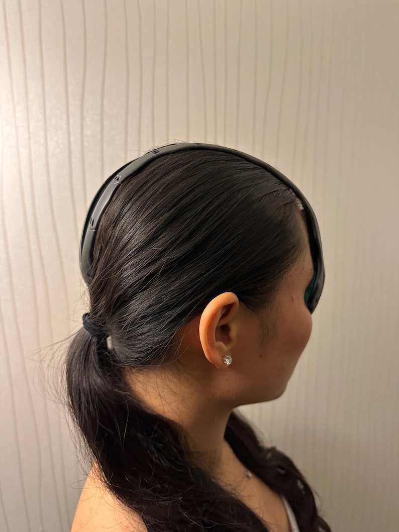 May include: A black headband worn on dark hair. The headband is smooth and curved, and it sits across the forehead. The hair is pulled back into a low ponytail. The person is wearing a small, clear stud earring.