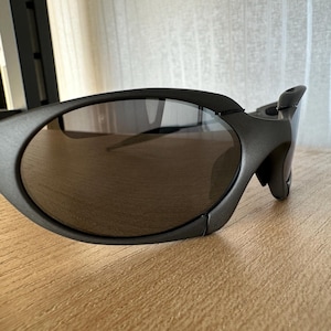 May include: Close-up of dark gray sunglasses with oval lenses. The sunglasses have a sleek, modern design with a curved frame and a distinctive temple detail. The lenses are a dark, reflective color.
