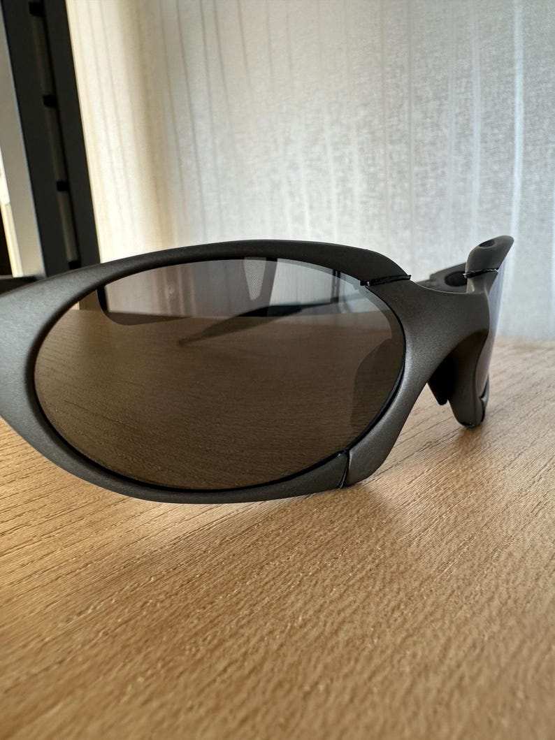 May include: Close-up of dark gray sunglasses with oval lenses. The sunglasses have a sleek, modern design with a curved frame and a distinctive temple detail. The lenses are a dark, reflective color.